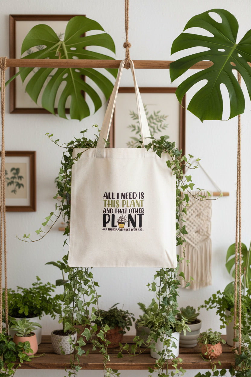 A white canvas tote bag hanging on a green plant hanger rack indoors. The bag has a humorous printed message that reads 'ALL I NEED IS THIS PLANT AND THAT OTHER PLANT' with a small potted plant graphic.